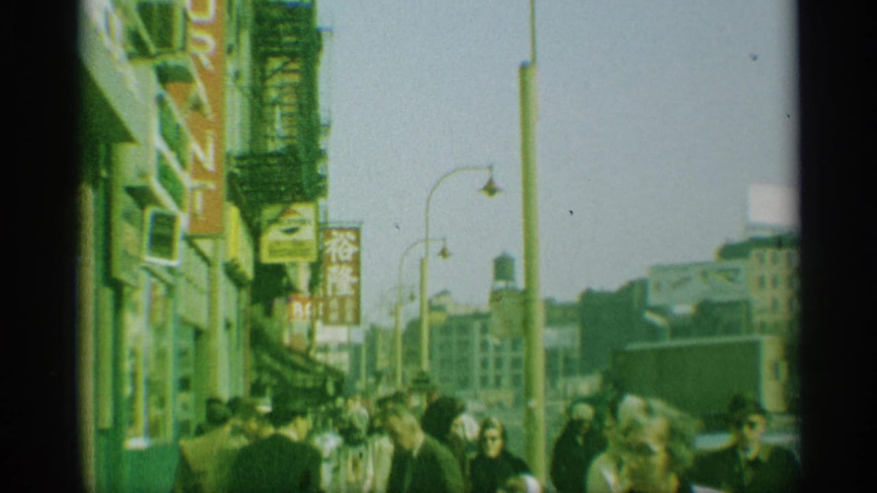 1959 New York street scene showing pedestrians, storefront signage, a tall lamppost, vehicles, and green-tinted 8mm texture.