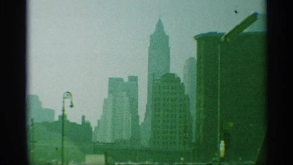 Late shot of the 1959 New York skyline, showing clustered towers, a foreground lamp, haze, and pronounced 8mm frame edges.