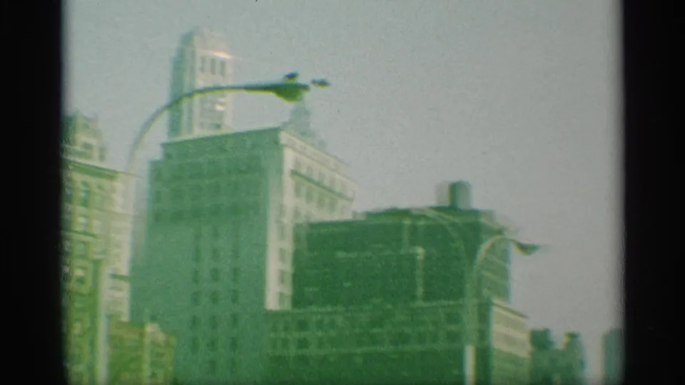 1959 New York street view with a curved lamp, mid-rise buildings, and washed pale sky in green-tinted 8mm footage.
