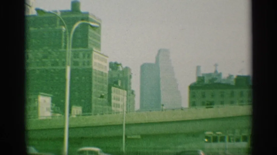1959 New York home-movie frame showing an elevated roadway, twin street lamps, and distant high-rises in haze.