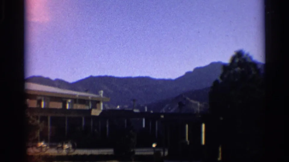 1959 New York home-movie frame with a flat-roof suburban house, shaded porch, utility lines, trees, and hills.