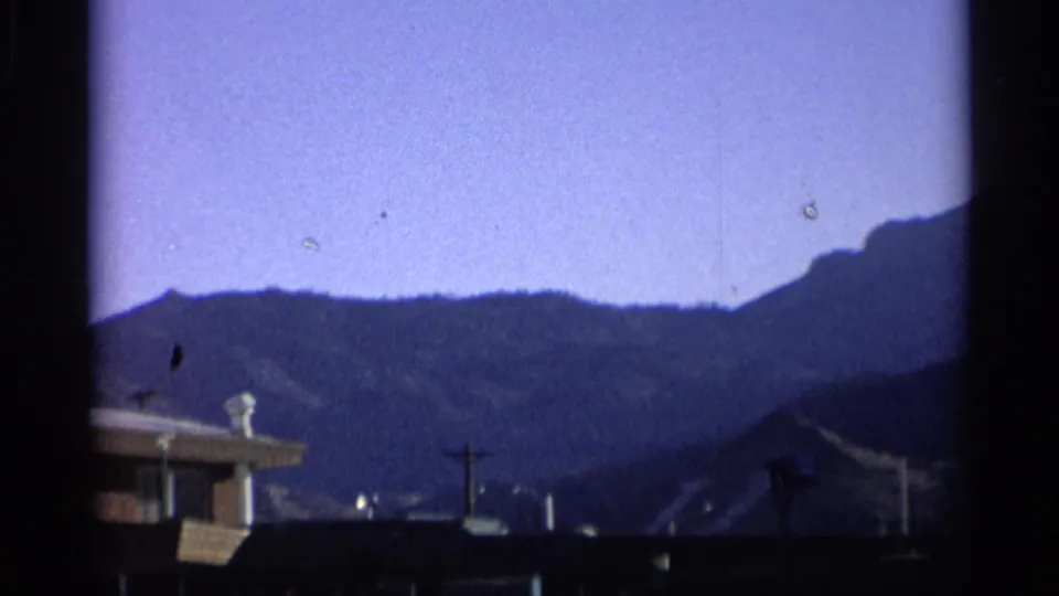1959 New York frame showing a house roofline, utility pole, hillside backdrop, and clear blue sky in faded 8mm color.
