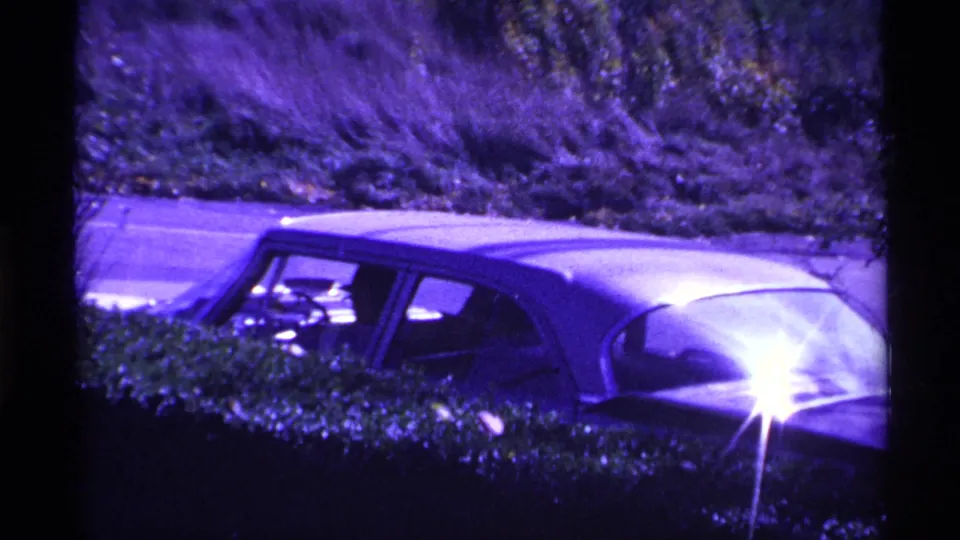 1969 Ohio home-movie frame showing a classic sedan near a hedge-lined roadside, with purple color shift and bright reflected light.