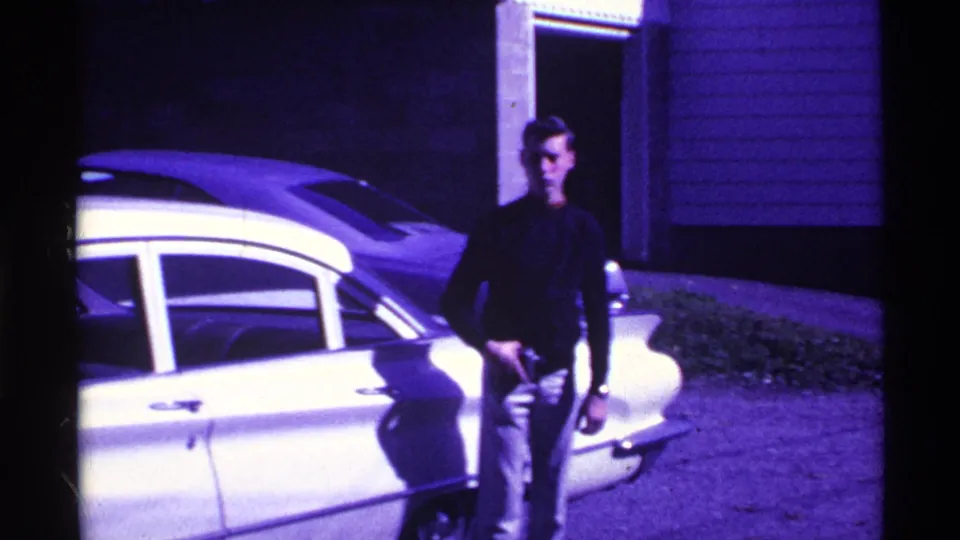 1969 Ohio 8mm frame showing a young man in a dark top and light pants beside a light-colored sedan near a building entrance.