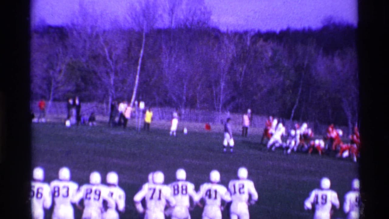 Football players line up on a grassy field with spectators and bare trees in the background.