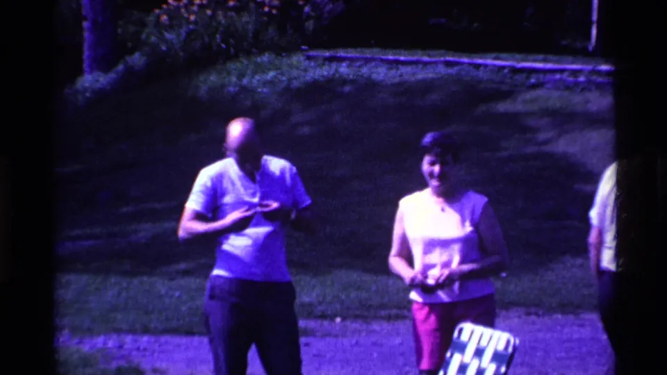 1969 Ohio home-movie frame of adult spectators near the sideline, including a bald man in a white shirt and a woman in a light top and pink pants.