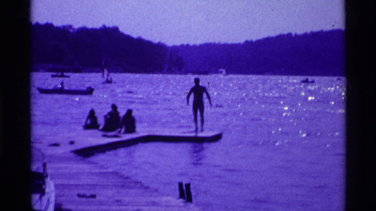 Person standing on a dock in a lake while others sit nearby, with boats and wooded shoreline in the background.
