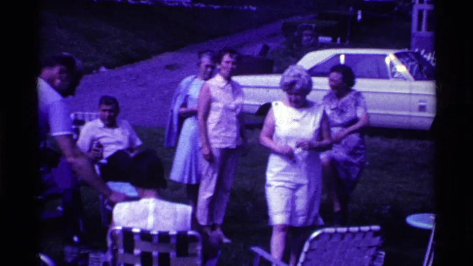 1969 Ohio home-movie frame showing adults in summer clothing gathered on a lawn near folding chairs and a white car.