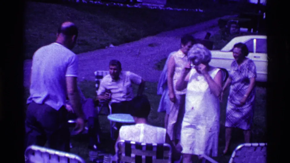 1969 Ohio backyard gathering frame with adults in summer attire near lawn chairs, a driveway, and a white car.