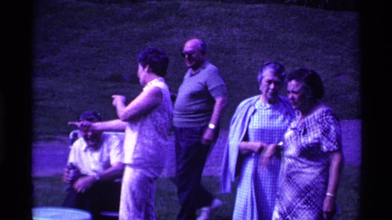 Adults in summer clothing stand and sit on a lawn near small outdoor tables.