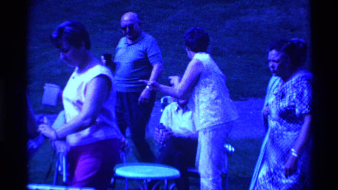 Several adults stand on a lawn, with a small table and chairs in the foreground.