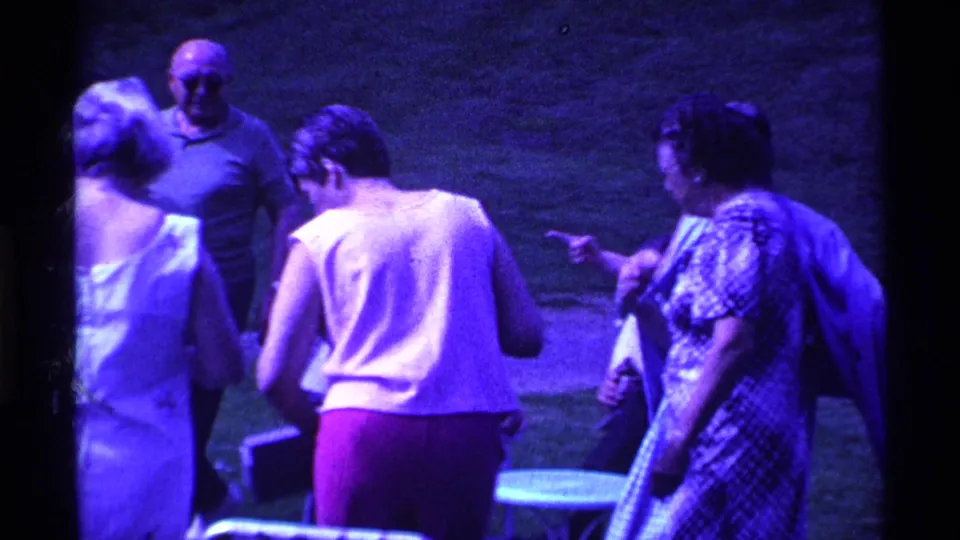 1969 Ohio home-movie frame showing adults in dresses and sleeveless tops gathered on grass near outdoor seating.