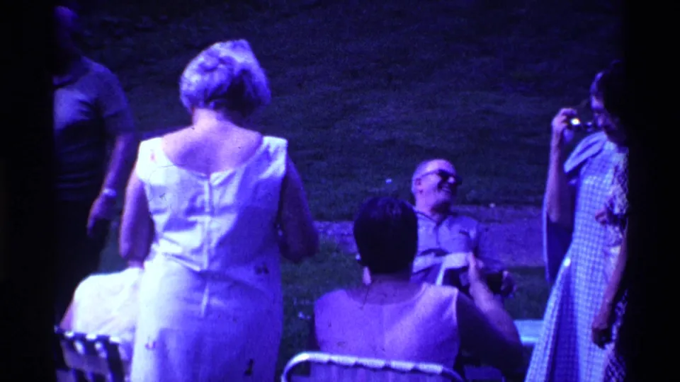 Late-1960s Ohio home-movie frame with adults socializing on a lawn and a woman at right appearing to hold a camera.