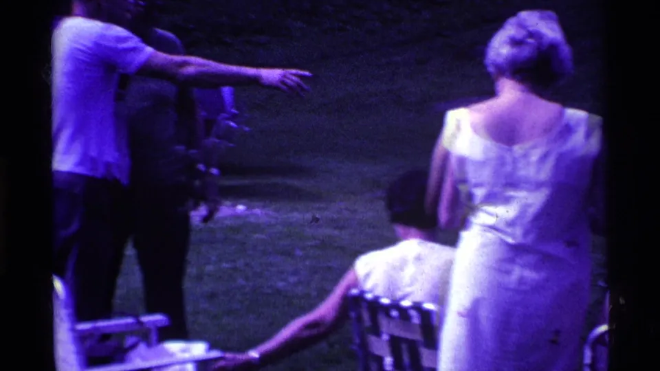 1969 Ohio backyard footage showing a man gesturing toward seated guests while adults gather on the grass in casual summer clothing.