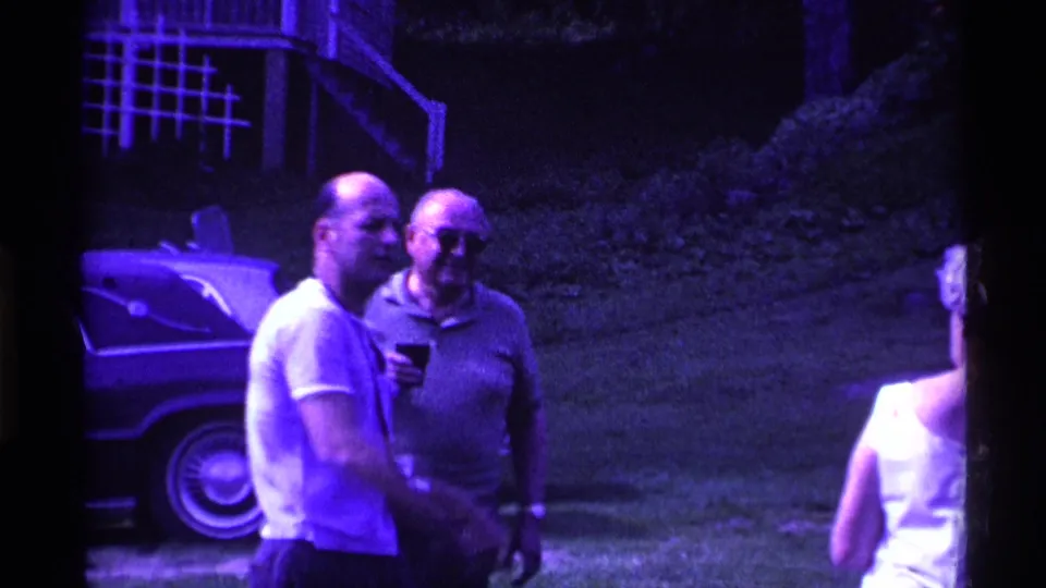 1969 Ohio home-movie frame with two men standing in conversation near a parked car at an outdoor backyard gathering.