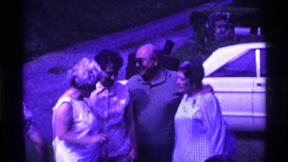 1969 Ohio 8mm frame showing four adults gathered in conversation near a light-colored car during a backyard social event.