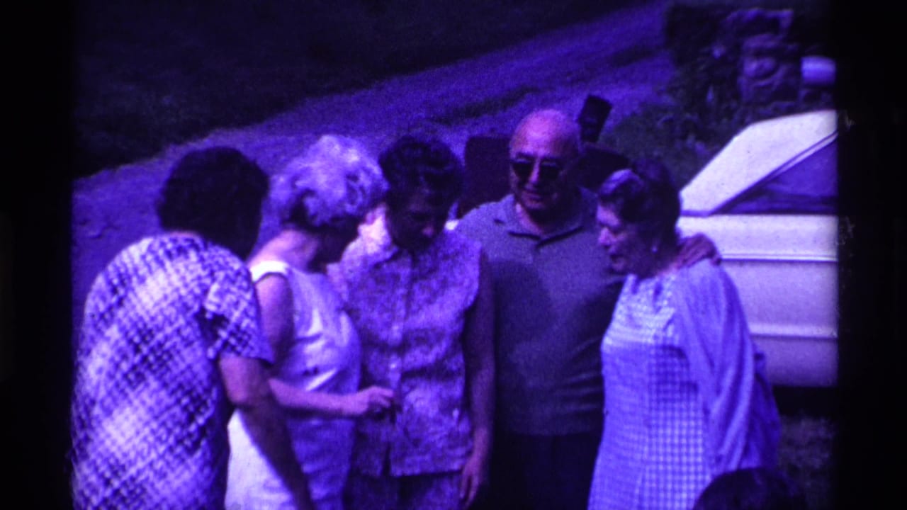 Adults gather outdoors beside a white car, with several women and a man in sunglasses facing the camera.