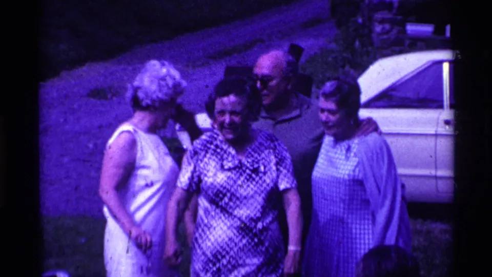 Adults in 1960s clothing smile and shift positions outdoors in Ohio, with the man in sunglasses standing near a white car.