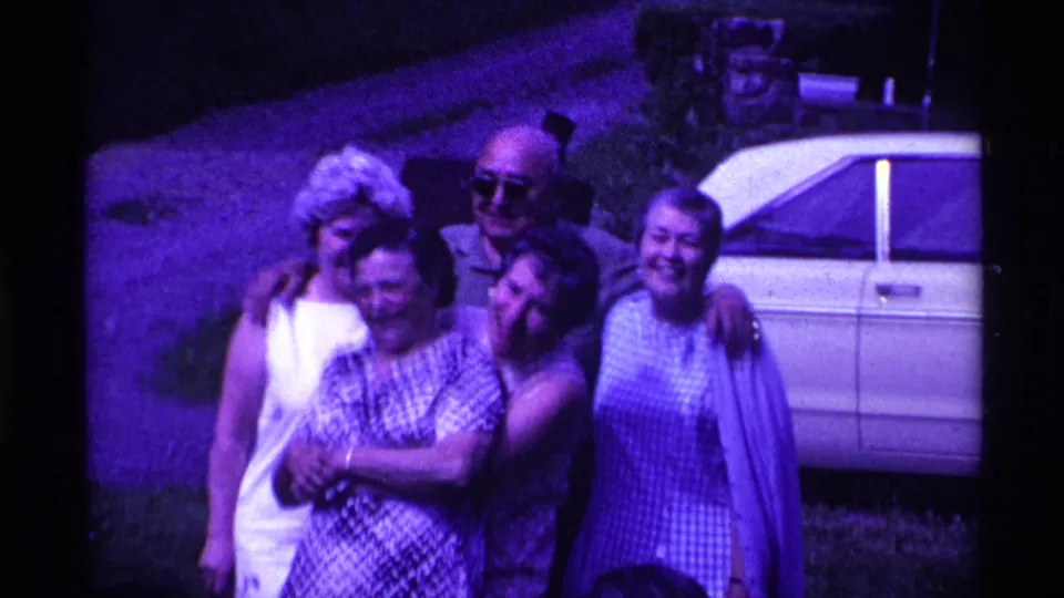 In this 1969 Ohio frame, adults lean together and embrace while posing for the camera beside a parked white car.
