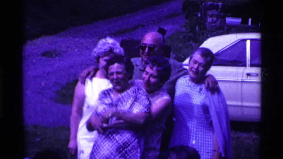 The 1969 Ohio home-movie frame shows five adults smiling in a close outdoor pose, with checked dresses and a white car behind them.