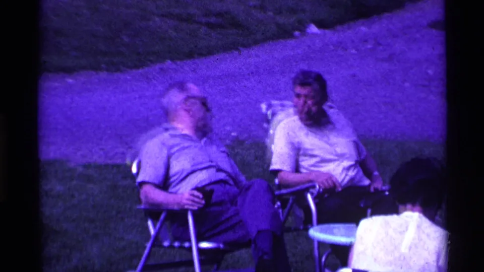 A home-movie frame shows seated men conversing in lawn chairs on an Ohio lawn, with a person seen from behind in the foreground.