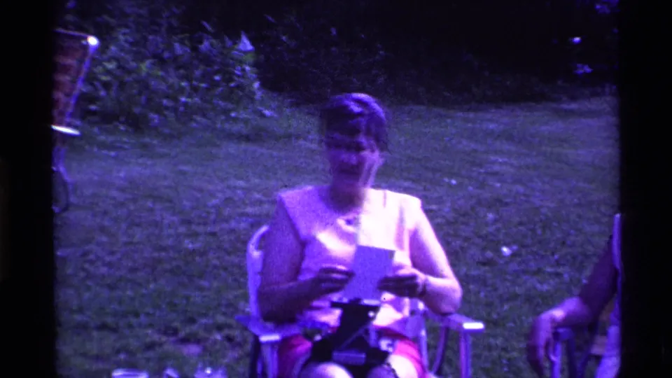 A woman at the 1969 Ohio gathering sits outdoors holding a card or letter, framed by grass and garden plants.