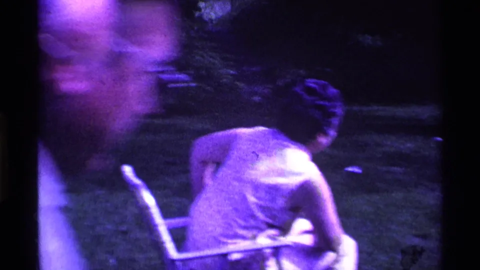 Purple-shifted 8mm frame with a blurred foreground figure and seated women in lawn chairs during an outdoor gathering.