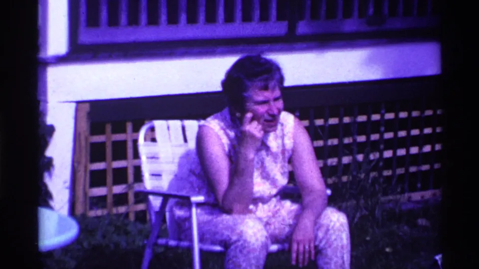 1969 Ohio home-movie frame showing an older woman seated by a lawn chair near a wooden deck railing, with purple color cast.