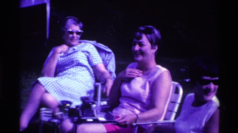 Three adult women sit together in folding lawn chairs on an Ohio lawn, smiling and conversing in 1969 8mm home-movie footage.