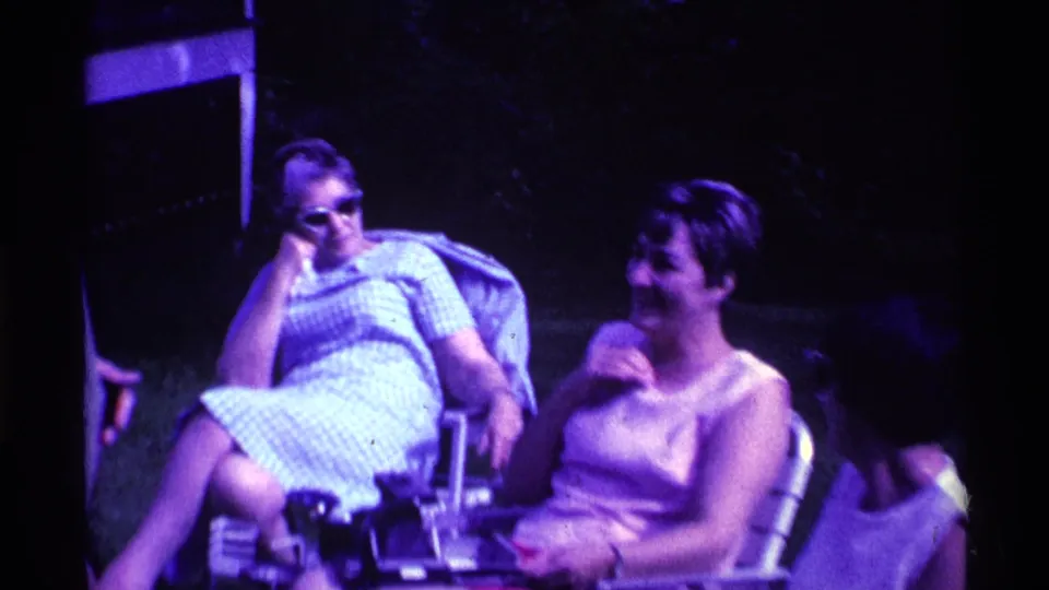 Two women remain seated in lawn chairs during the backyard gathering, with a partially visible person at the frame edge and a porch behind them.
