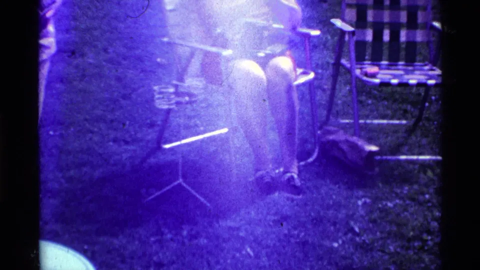 A bright, overexposed 8mm frame shows a woman seated on the grass-side lawn chair area, with other chairs nearby and heavy purple film flare.