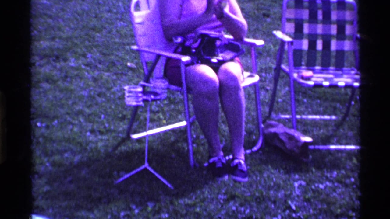 In Ohio 1969 home-movie footage, a woman sits in a lawn chair on grass with a plaid folding chair and small stand nearby.