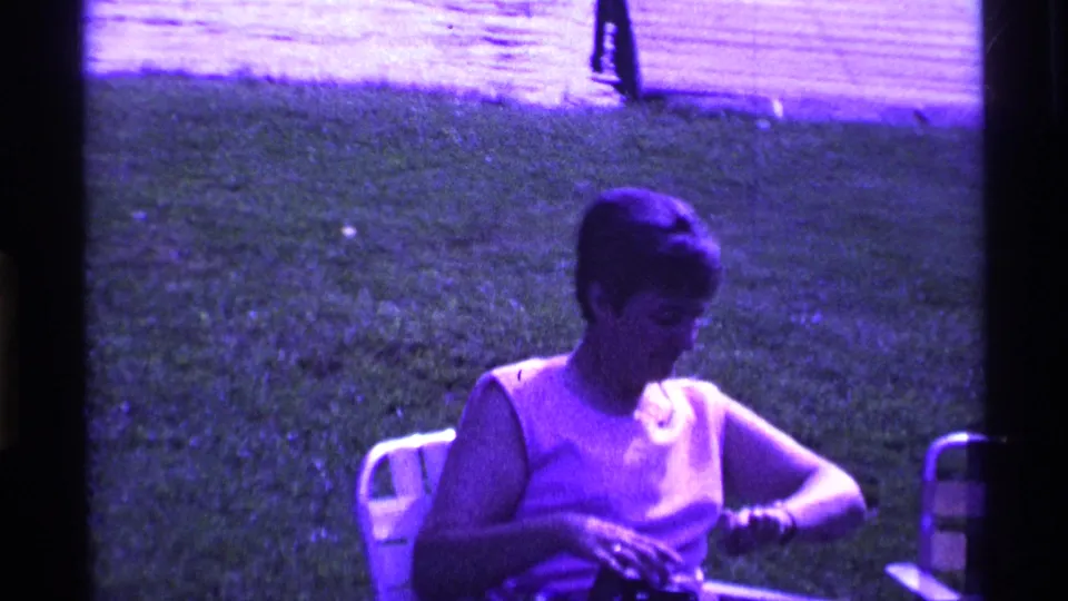 A closer 8mm frame shows the seated woman adjusting the camera or case while leaning slightly forward in the grassy yard.