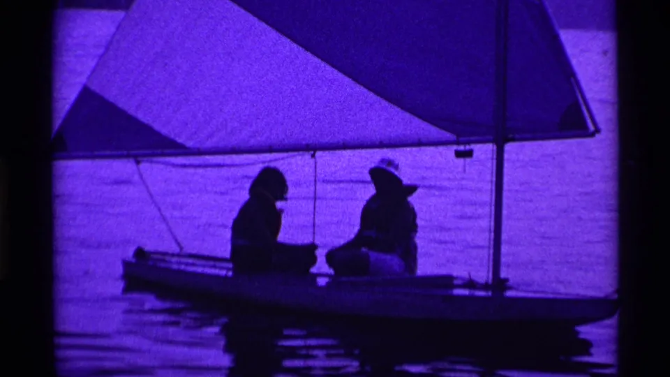 Two silhouetted people appear to converse aboard a small sailboat on a purple-tinted Ohio lake in 1969 footage.