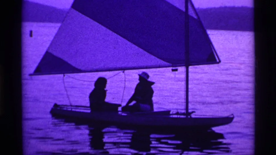 A small sailboat with two seated figures crosses a purple-tinted lake in archival 1969 Ohio 8mm footage.