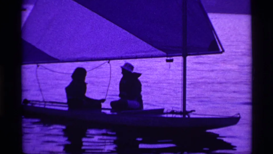 Two people sit in silhouette on a small sailboat, framed by mast, sail, and rippled Ohio lake water in 1969 footage.