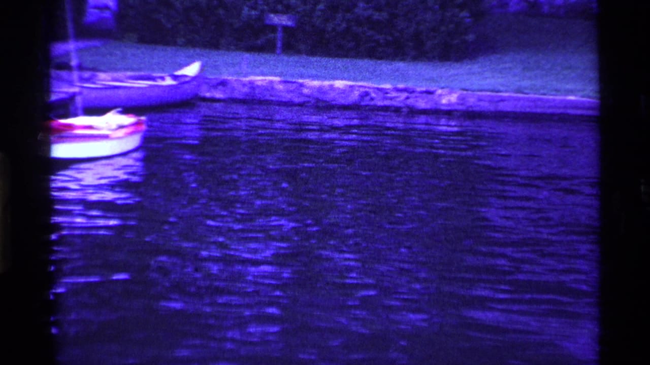 1969 Ohio lakeside home-movie frame with moored small sailboats, shoreline grass, trees, and purple-blue water reflections.
