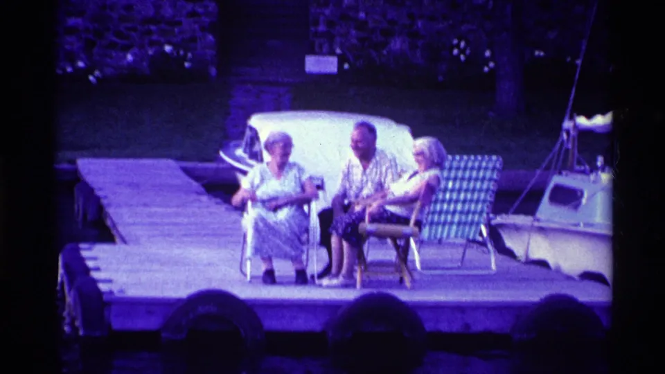 1969 Ohio 8mm frame of three adults seated in lawn chairs on a dock, with a small sailboat, water, and shaded shoreline behind them.