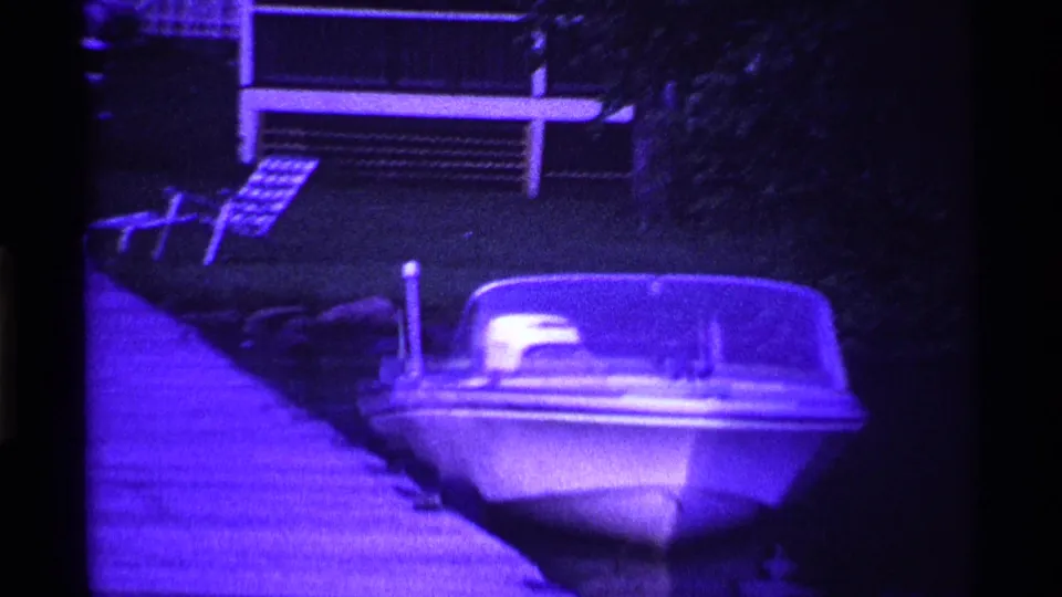 1969 Ohio home-movie frame with a moored motorboat, dock, lawn, and house exterior under a strong purple cast.