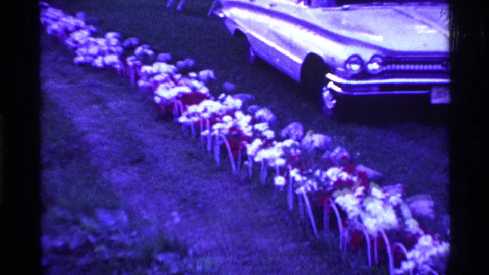 The pan ends on artificial flowers beside the front end of a light-colored classic American car in an Ohio yard.