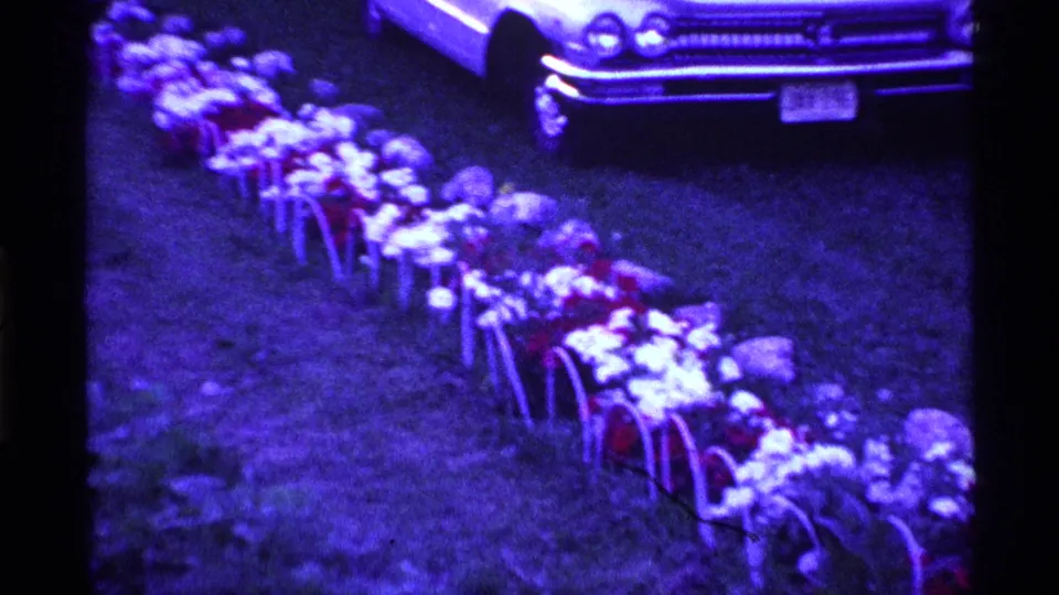 1969 Ohio 8mm frame showing a white vintage sedan near a grave-side row of white and red flowers in metal holders.