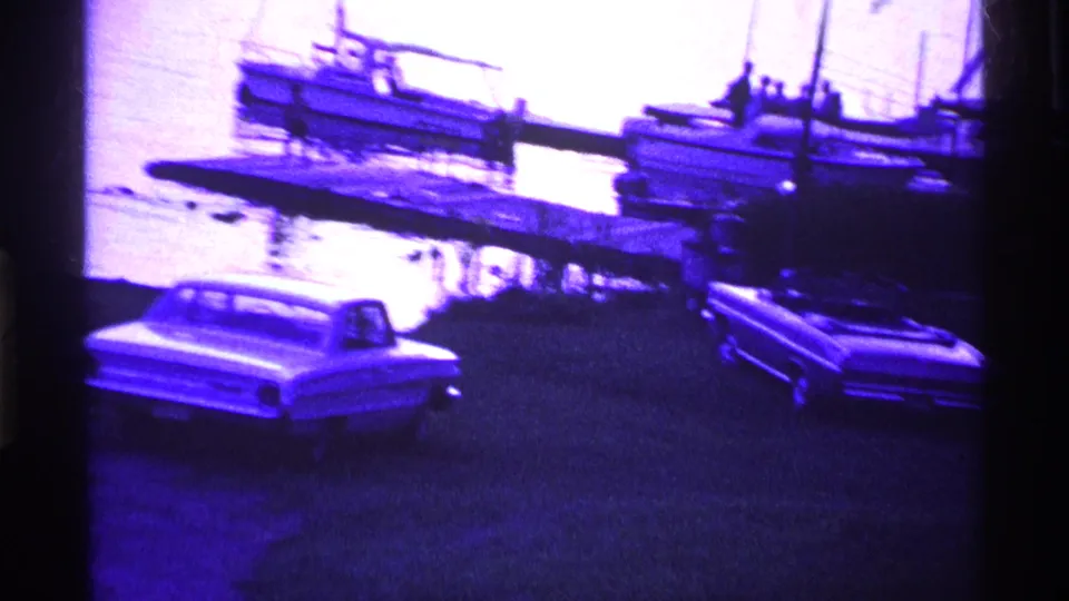 1969 Ohio 8mm frame showing parked vintage cars near a dock where small boats are tied on calm water.