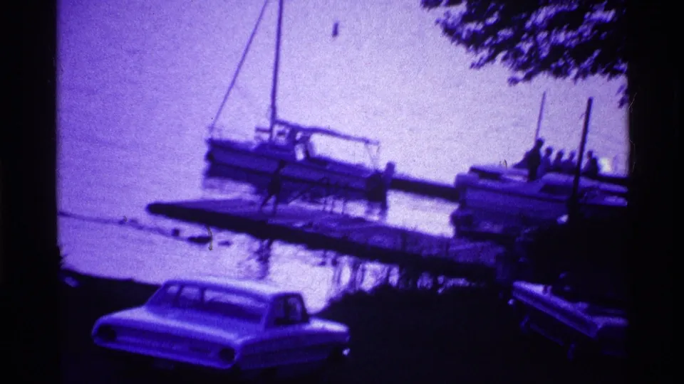 Purple-tinted Ohio home-movie frame with small boats moored at a dock and a vintage car in the foreground.