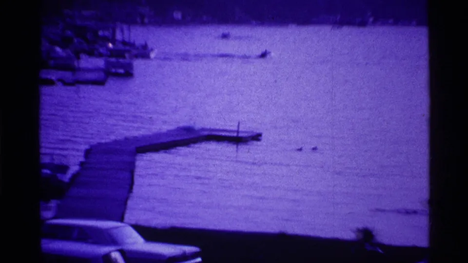 Purple-toned 1969 Ohio home-movie frame showing a lakeside dock, calm water, marina boats, and part of a parked car.