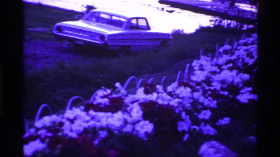 1969 Ohio home-movie frame showing a pale classic sedan behind white and red flowers in a grassy yard.