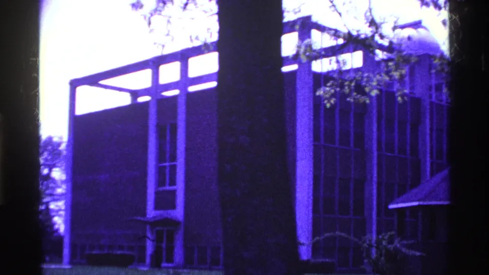1969 Ohio frame showing the corner of a taller brick institutional building with vertical window bays behind a tree.