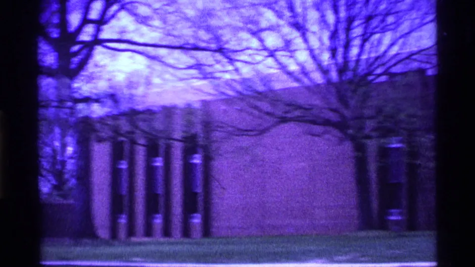 1969 Ohio 8mm frame showing a purple-tinted modernist wall, vertical window openings, lawn, and bare tree branches.