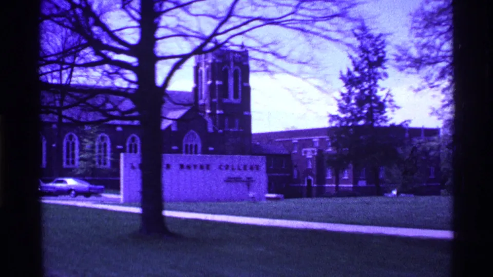 1969 Ohio campus view with a large college sign, brick tower building, parked car, lawn, sidewalk, and bare trees in purple-tinted 8mm footage.