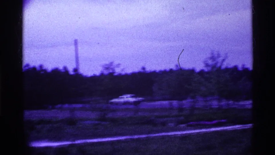 Purple-tinted 1969 Ohio roadside view with a distant car, tree line, open land, and a tall vertical tower.