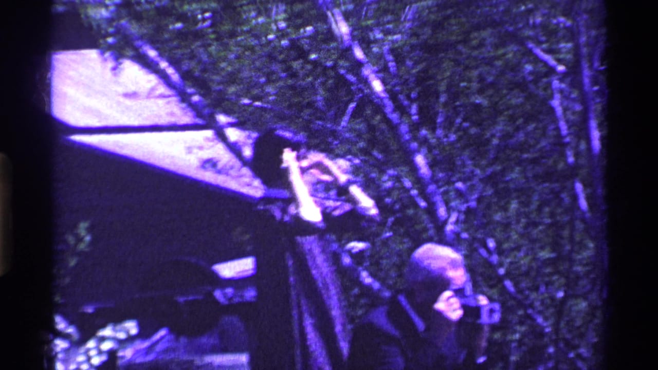 Older man filming outdoors beside trees and a house roofline.